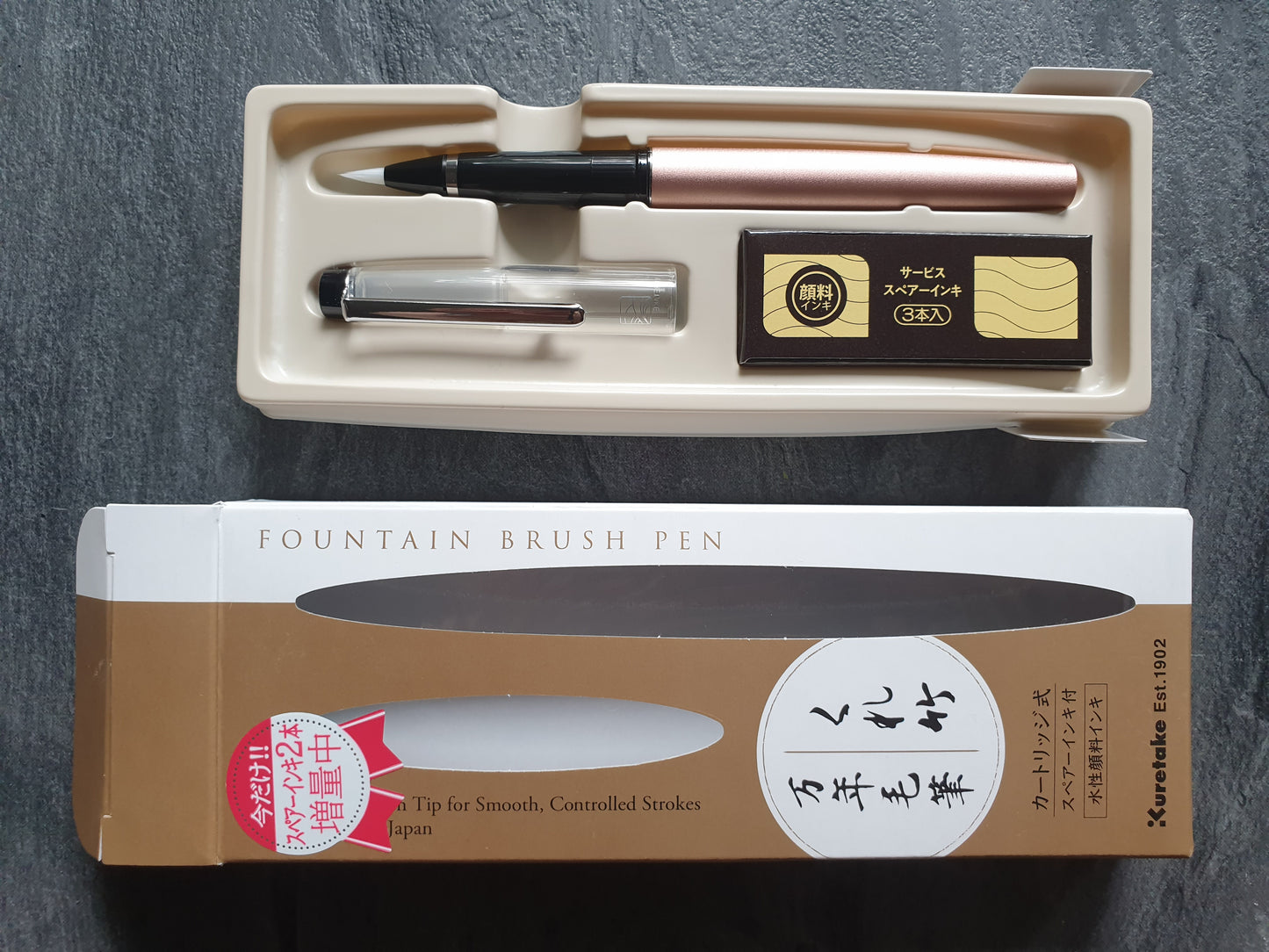 Ein rosegoldfarbener DT120 Pinselstift liegt im Plastik-Tray, die Pappschachtel liegt daneben. Im Tray steckt auch eine Packung mit 3 Tintenpatronen.
