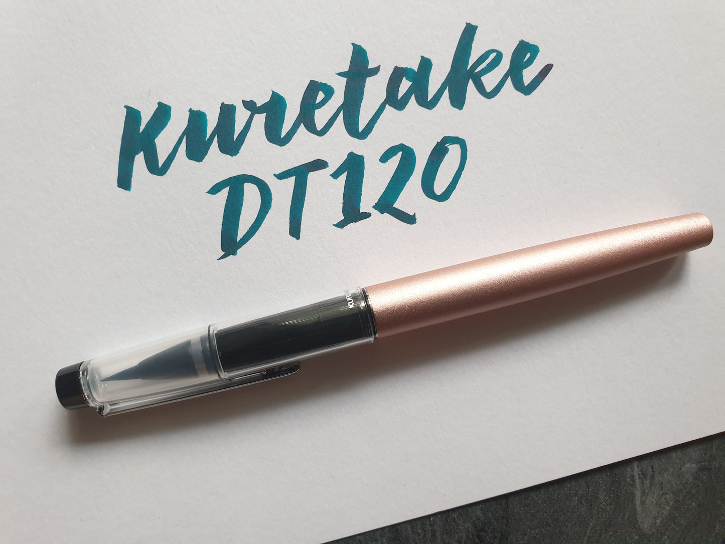 Ein rosegoldfarbener Pinselstift liegt neben dem Text, der damit geschrieben wurde.