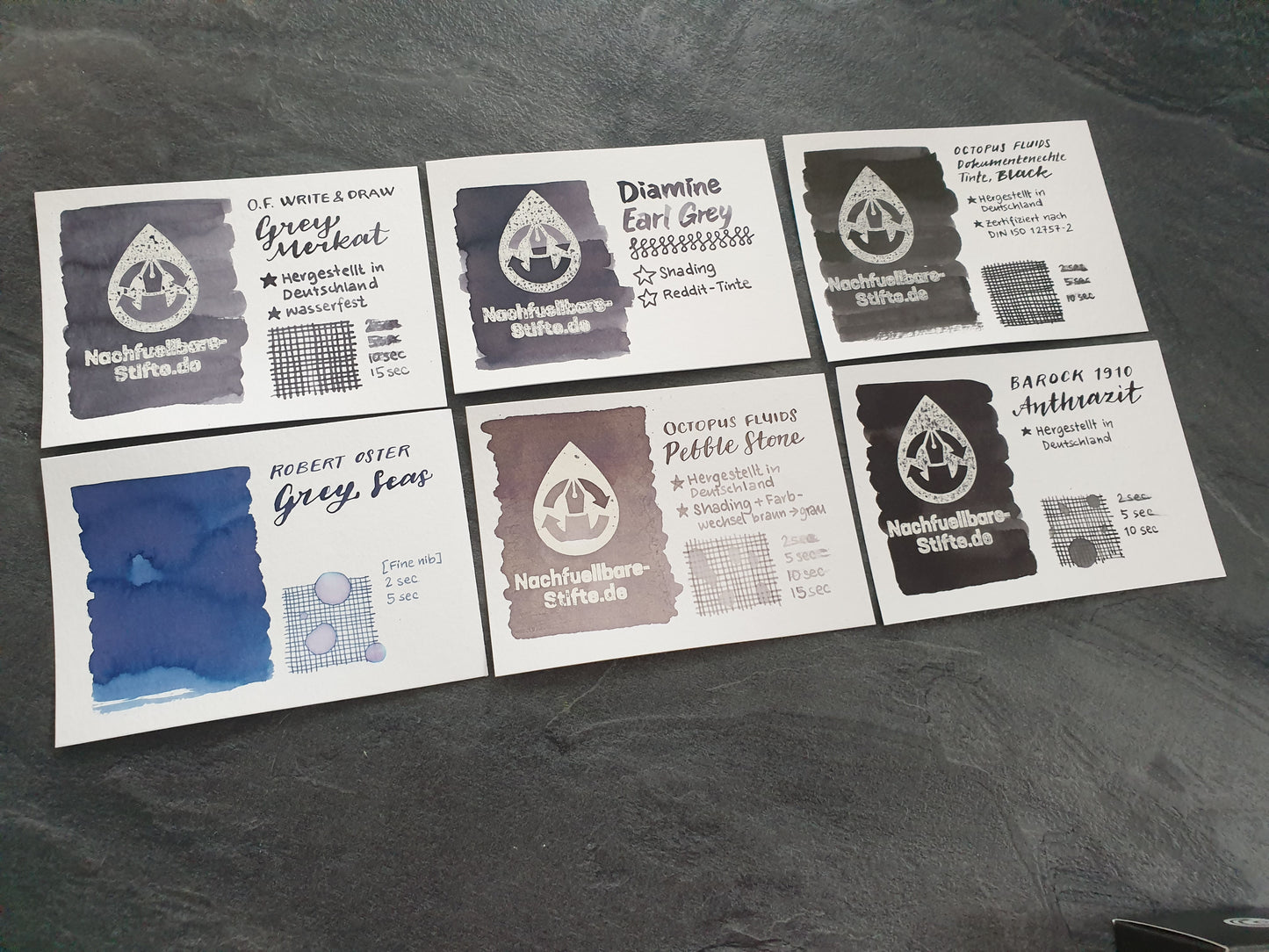 Vergleichsfoto mit Tintenswatches der Füllertinten: Octopus Fluids Write & Draw Grey Merkat, Diamine Earl Grey, Octopus Fluids Dokumentenechte Tinte Black, Robert Oster Grey Seas, Octopus Fluids Pebble Stone, Barock 1910 Anthrazit